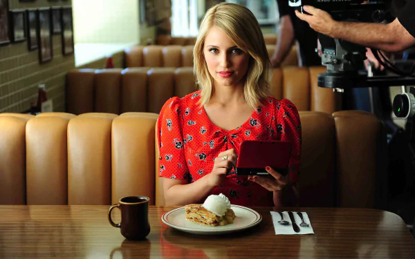 Dianna Agron sits at a diner table, holding a handheld gaming device, with a plate of food and a coffee cup in front of her. The vibrant scene serves as an engaging HD desktop wallpaper.