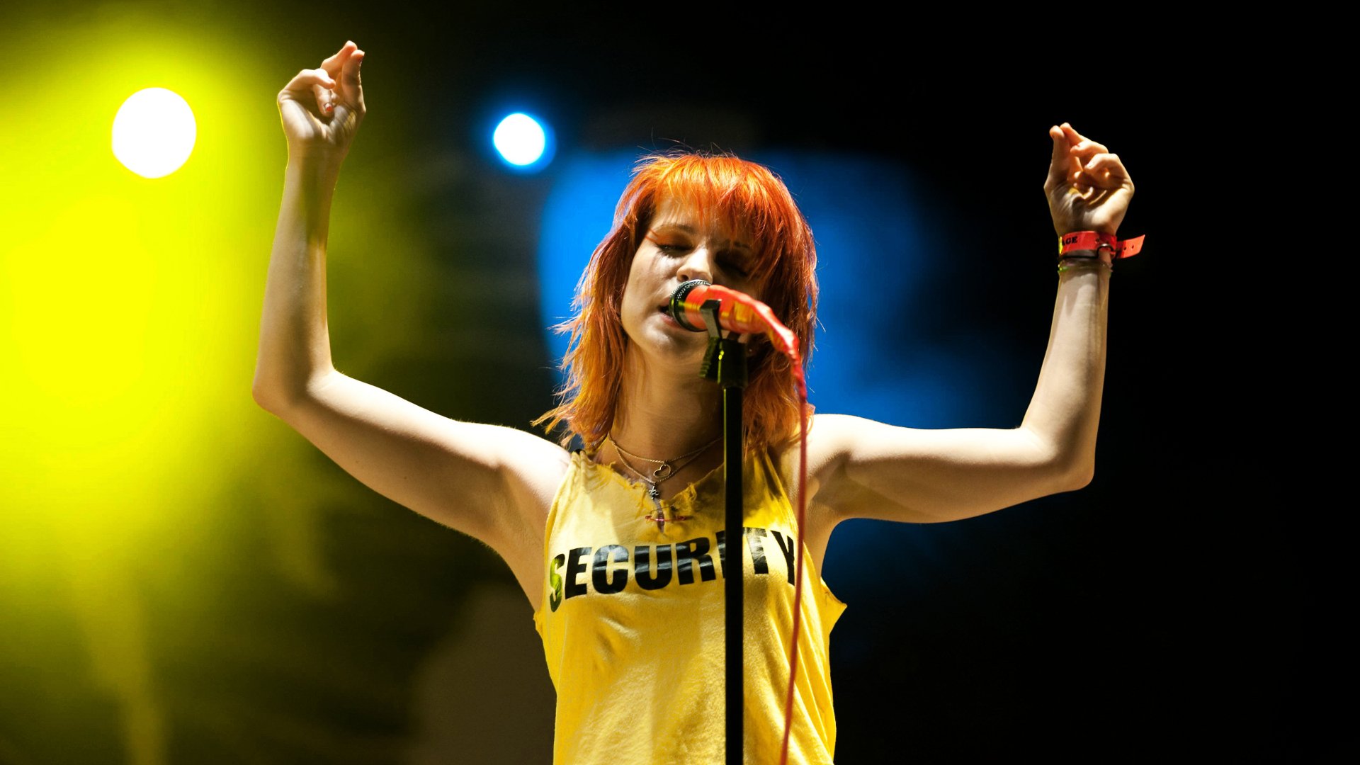 HD PC wallpaper of a female rock singer with bright orange hair in a yellow tank top, arms raised, singing into a mic under vibrant stage spotlights.