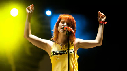 HD PC wallpaper of a female rock singer with bright orange hair in a yellow tank top, arms raised, singing into a mic under vibrant stage spotlights.