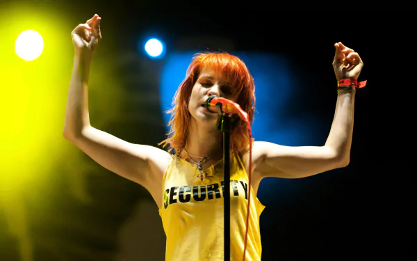 HD PC wallpaper of a female rock singer with bright orange hair in a yellow tank top, arms raised, singing into a mic under vibrant stage spotlights.