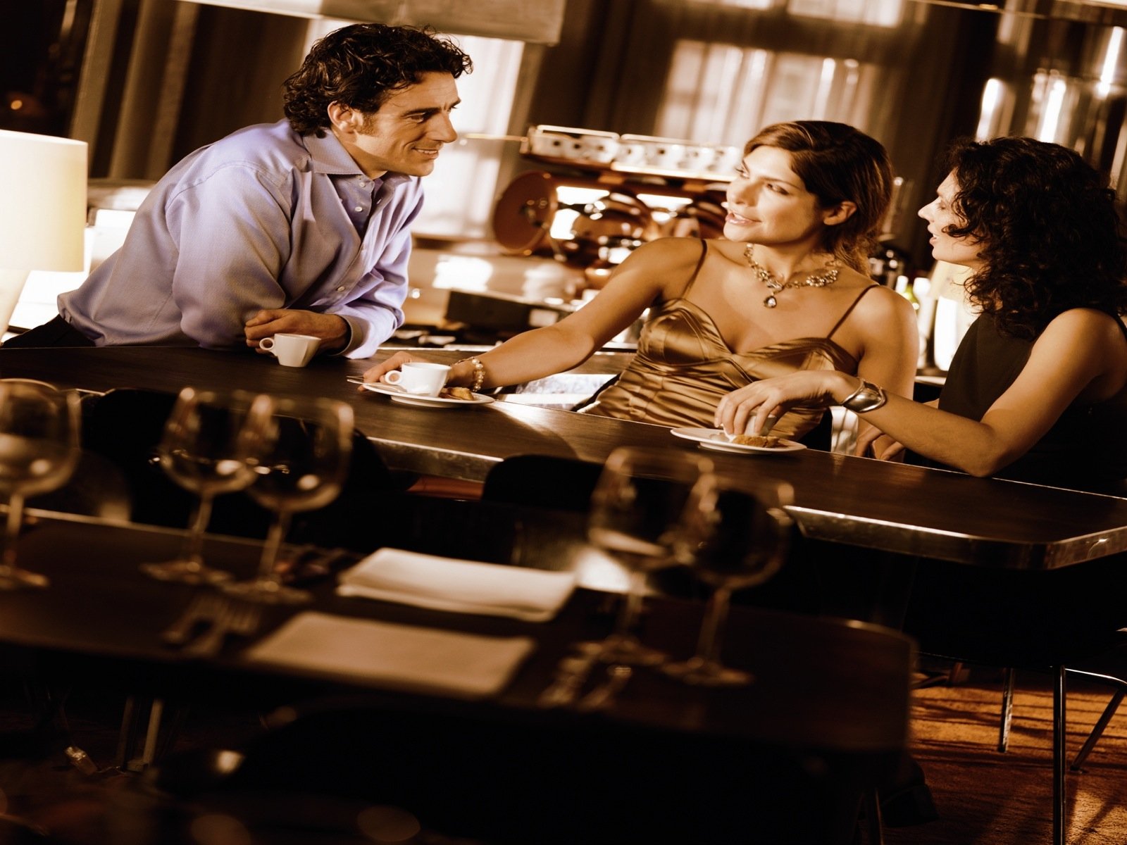 HD PC desktop wallpaper: warm photography of three well-dressed people laughing and chatting at a dimly lit bar, glasses and tableware in the foreground.