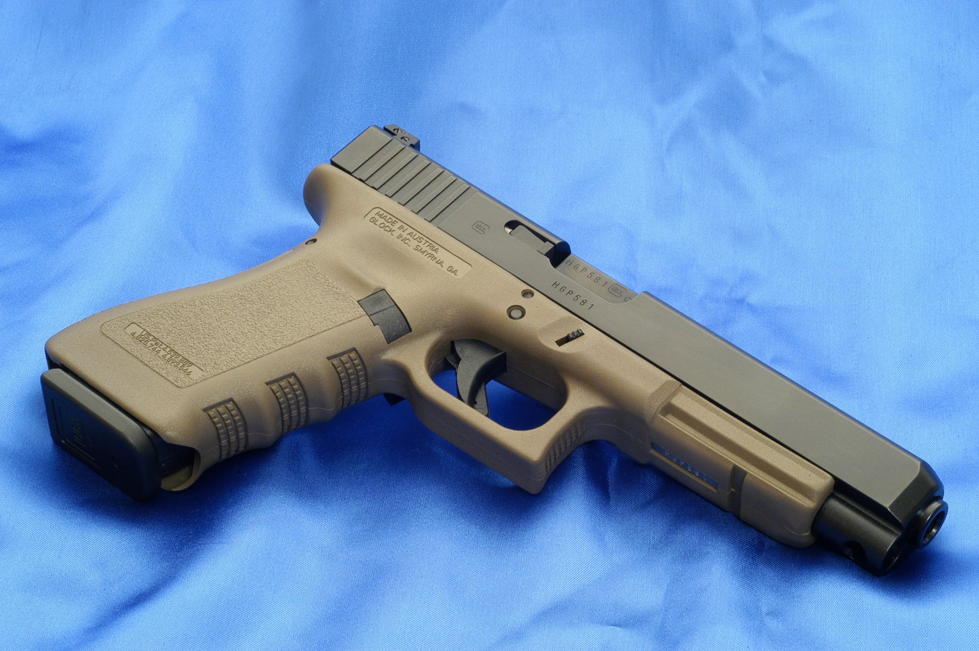 HD desktop wallpaper featuring a Glock pistol resting on a blue background. The firearm has a tan grip and a black slide, showcasing its detailed construction.