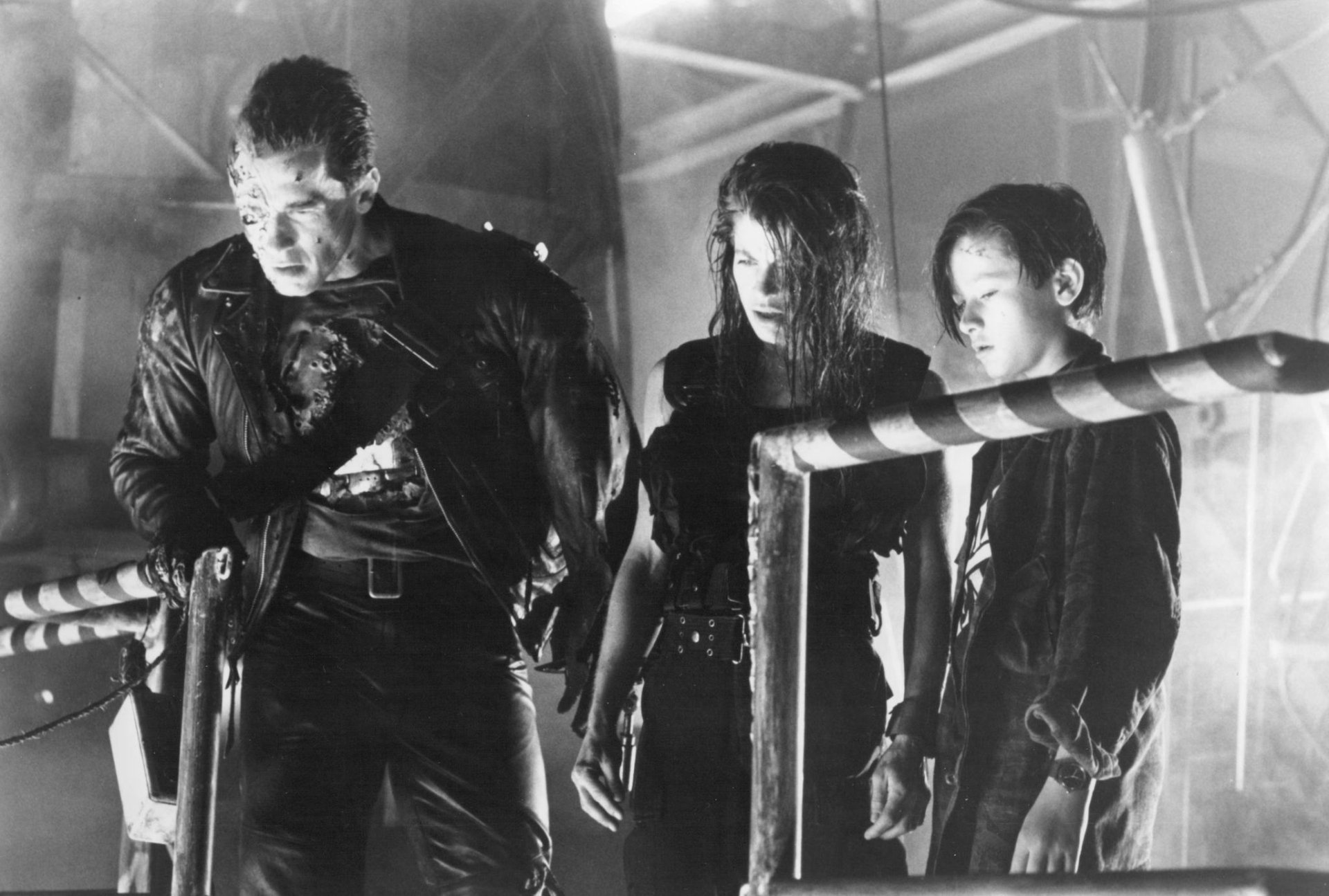 A dramatic scene from *Terminator 2: Judgment Day*, featuring Arnold Schwarzenegger as the Terminator, Linda Hamilton as Sarah Connor, and Edward Furlong as John Connor in a tense moment.