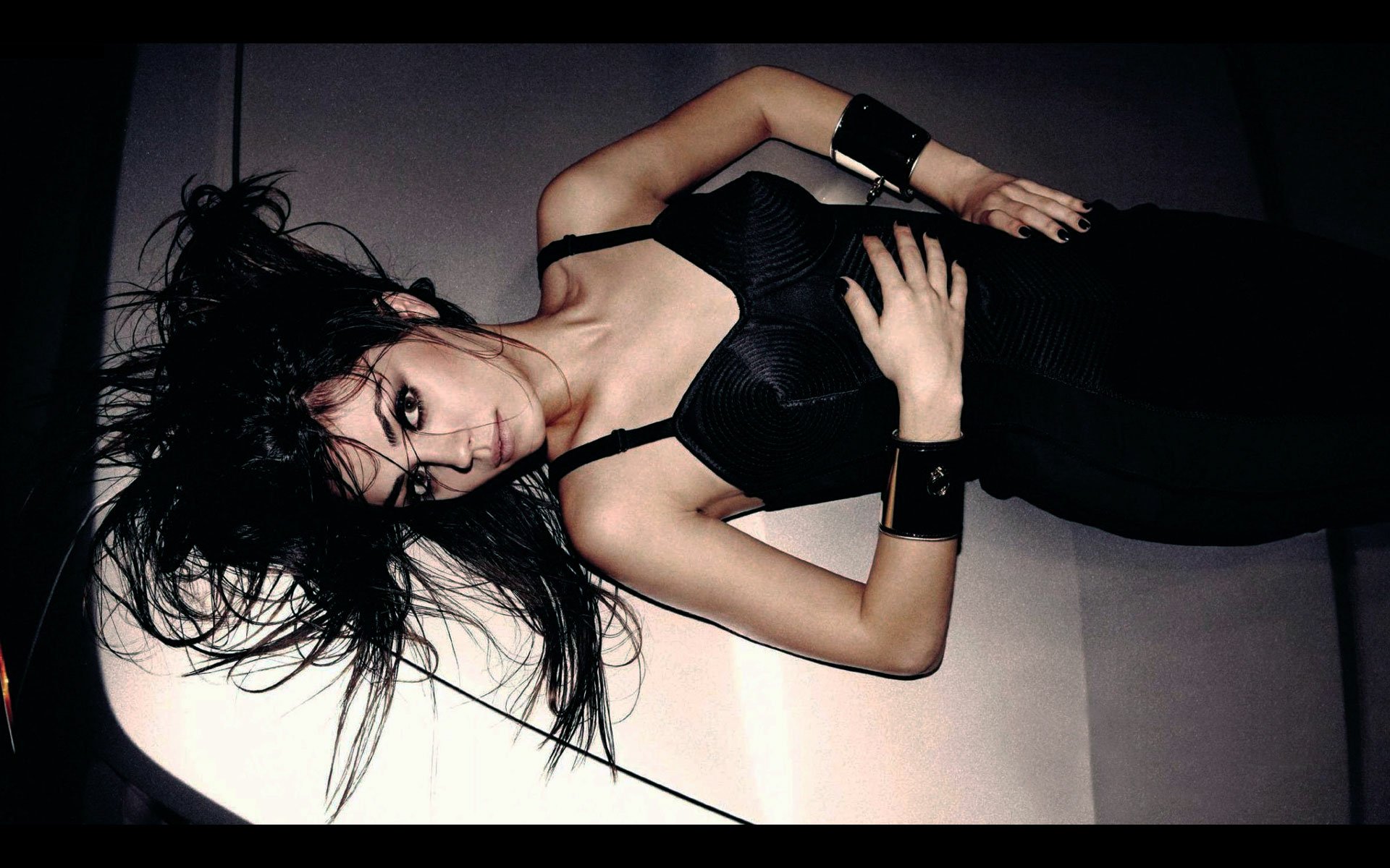 HD desktop wallpaper featuring celebrity Mila Kunis lying down, dressed in black with tousled hair and a confident pose against a minimalist background.