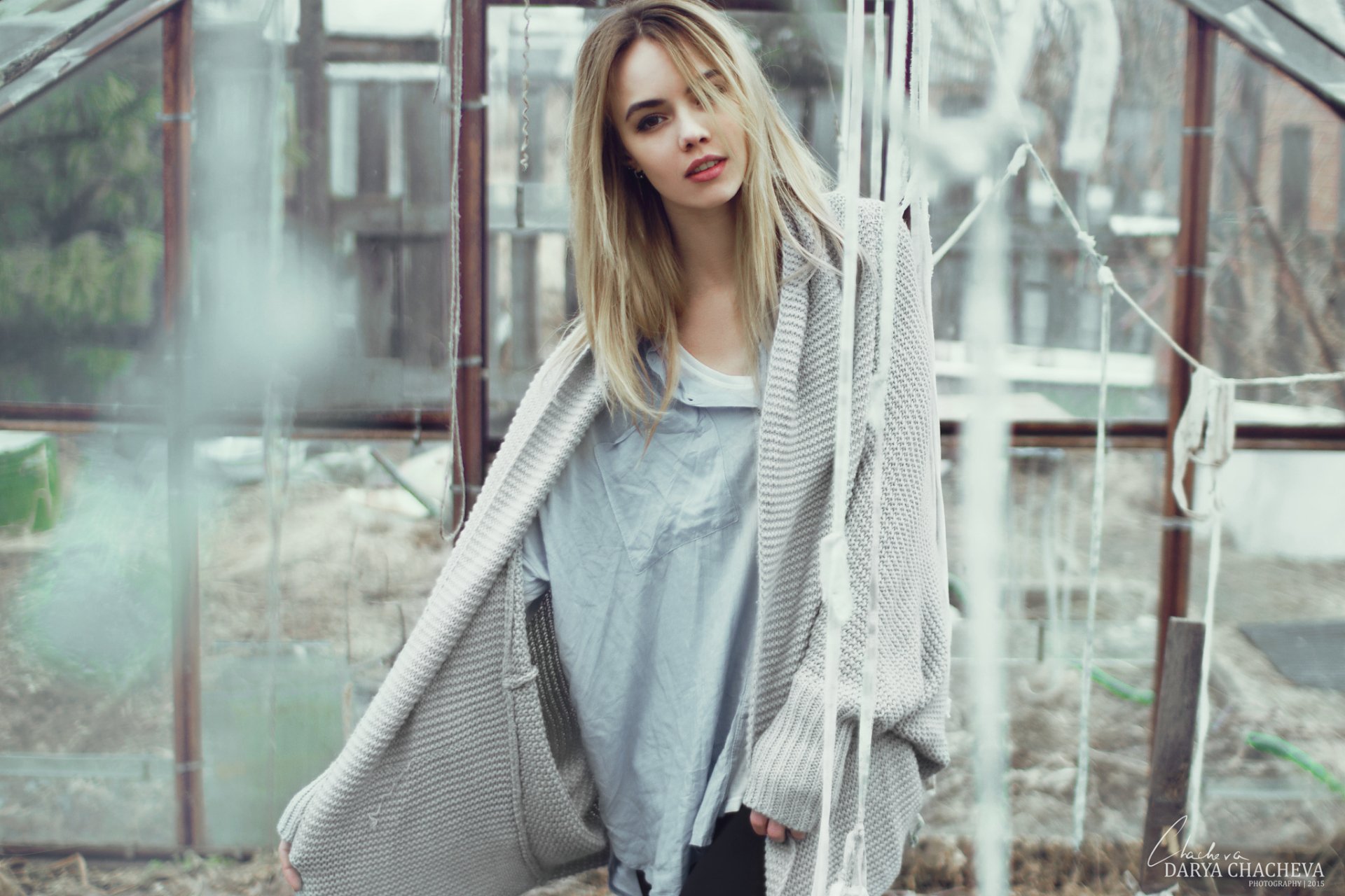 An HD desktop wallpaper featuring a woman with long blonde hair, wearing an oversized gray cardigan, standing in a rustic outdoor setting with a glasshouse in the background.