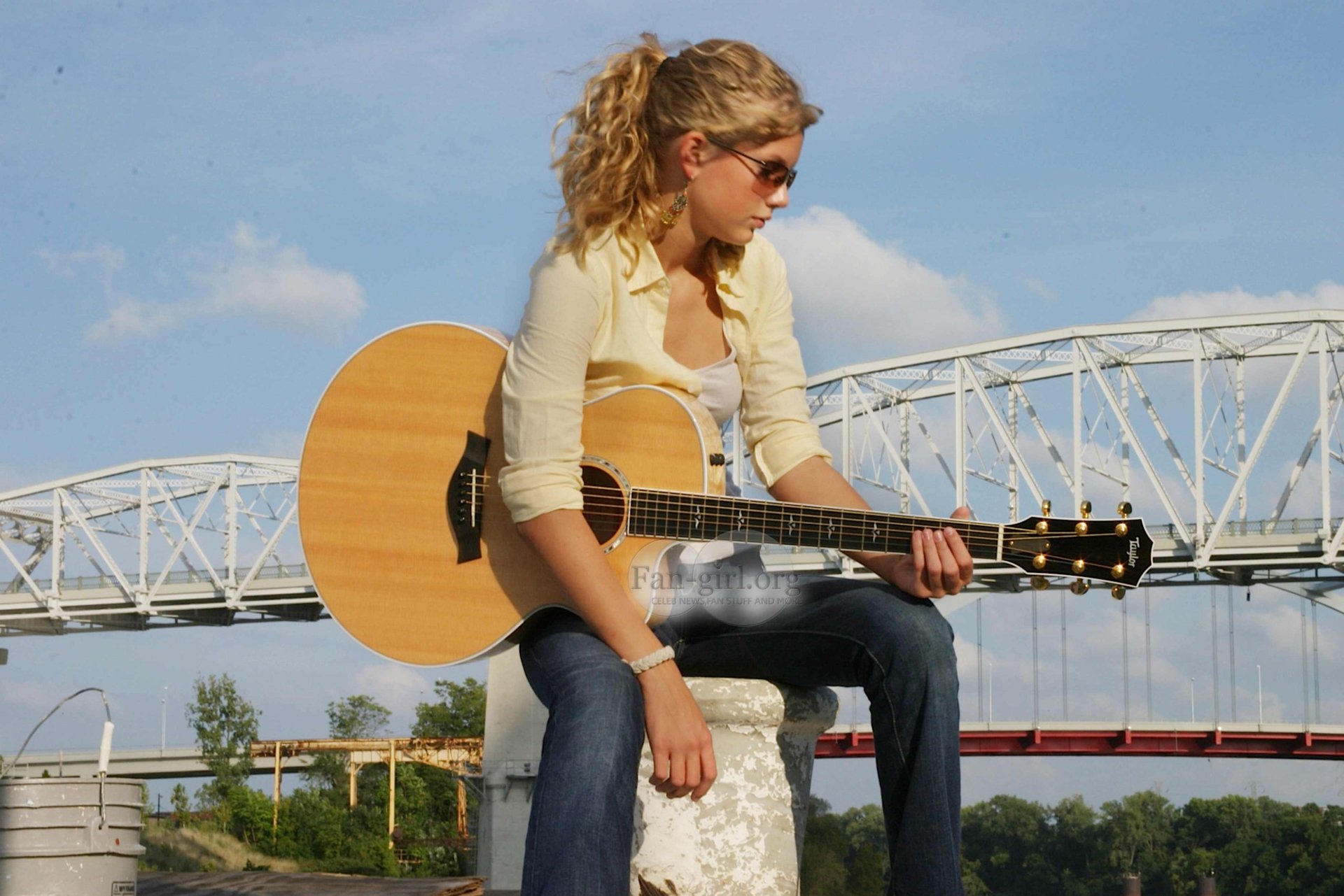 Taylor Swift playing an acoustic guitar outdoors with a bridge in the background, depicted in a vibrant 4K Ultra HD music-themed desktop wallpaper.