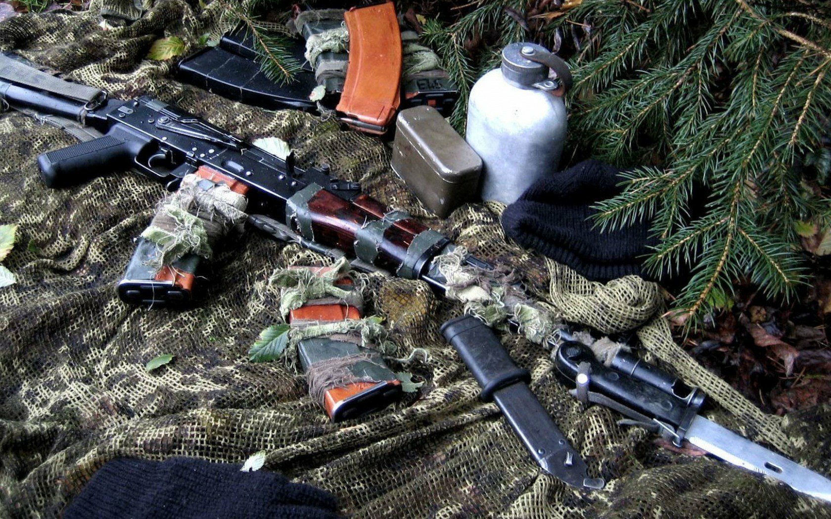 A detailed HD desktop wallpaper featuring an AK-47 surrounded by ammunition and supplies, set against a backdrop of camouflaged fabric and natural foliage.