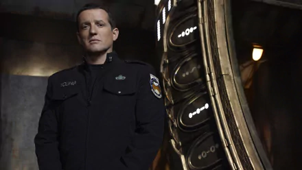 HD PC desktop wallpaper: stern officer from the TV show Stargate Universe stands beside a glowing stargate ring in a dim, industrial ship corridor.