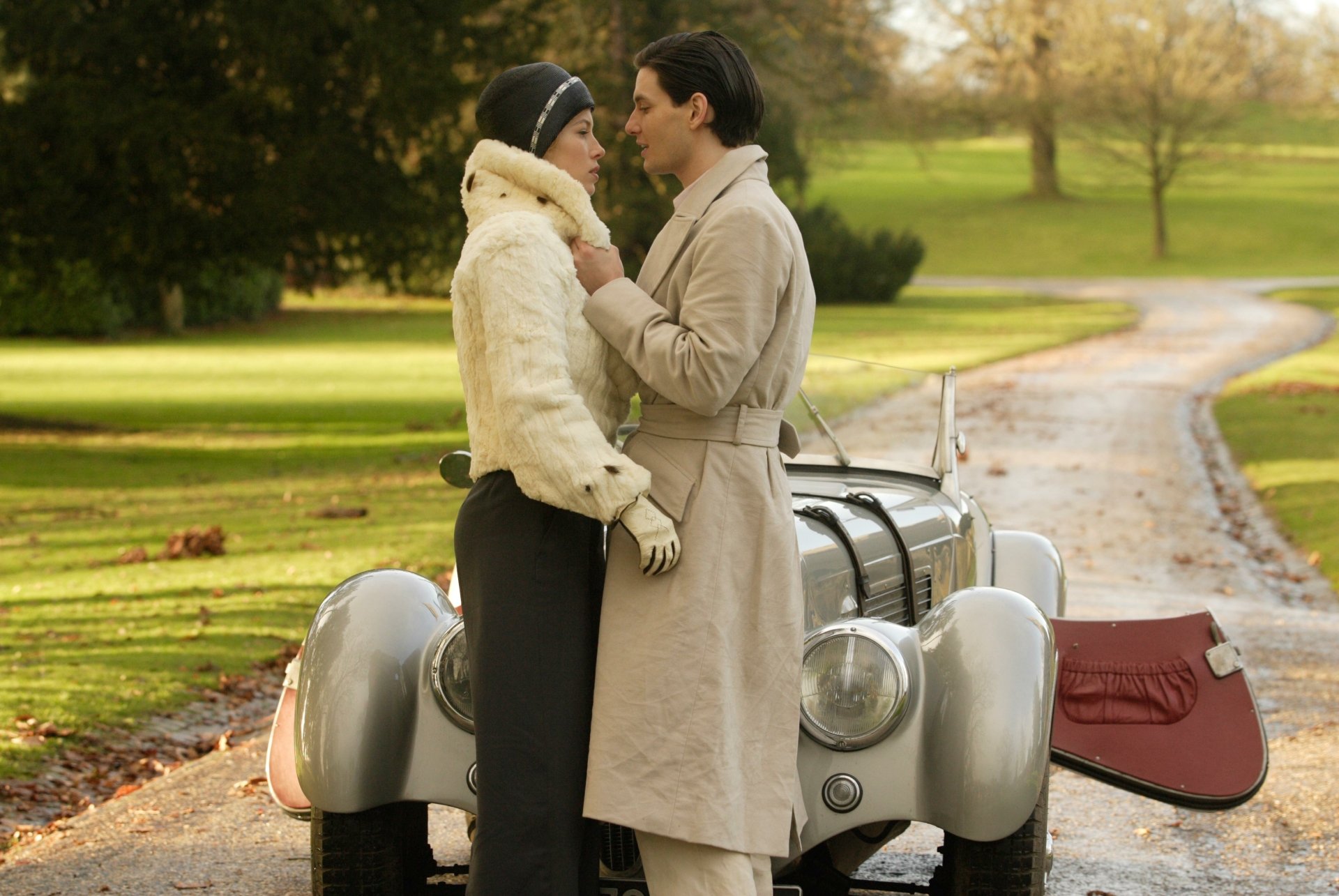 Period-romance scene: couple in 1930s attire embrace beside a vintage car on a leafy drive — 4K Ultra HD PC desktop wallpaper inspired by the film Easy Virtue.
