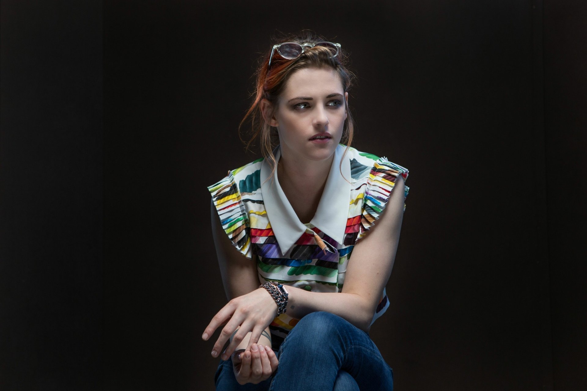 A contemplative Kristen Stewart, an American actress, is captured in a stylish, colorful top, sitting against a dark background, creating an engaging HD wallpaper.