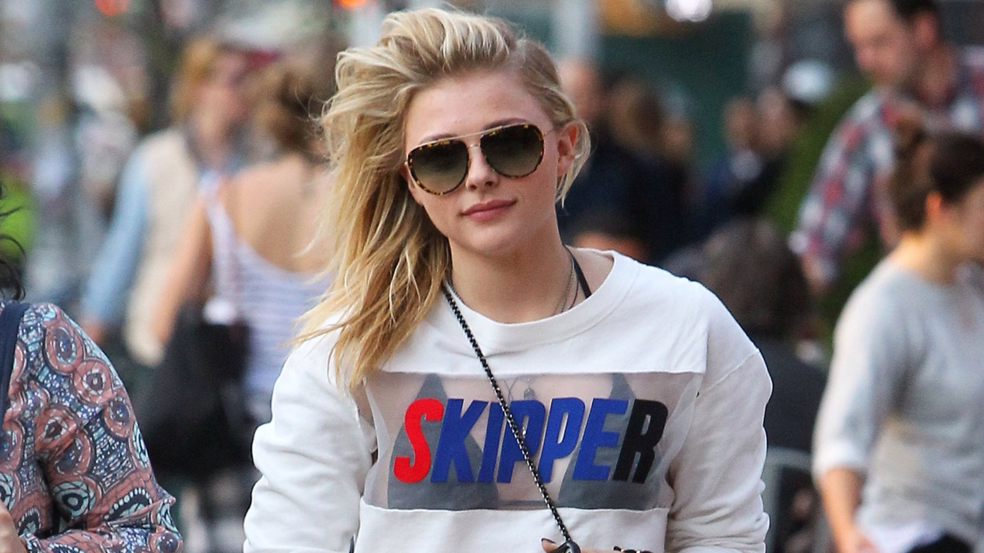 Chloë Grace Moretz walks confidently down a bustling street, wearing stylish sunglasses and a graphic sweatshirt, captured in a vibrant HD desktop wallpaper.