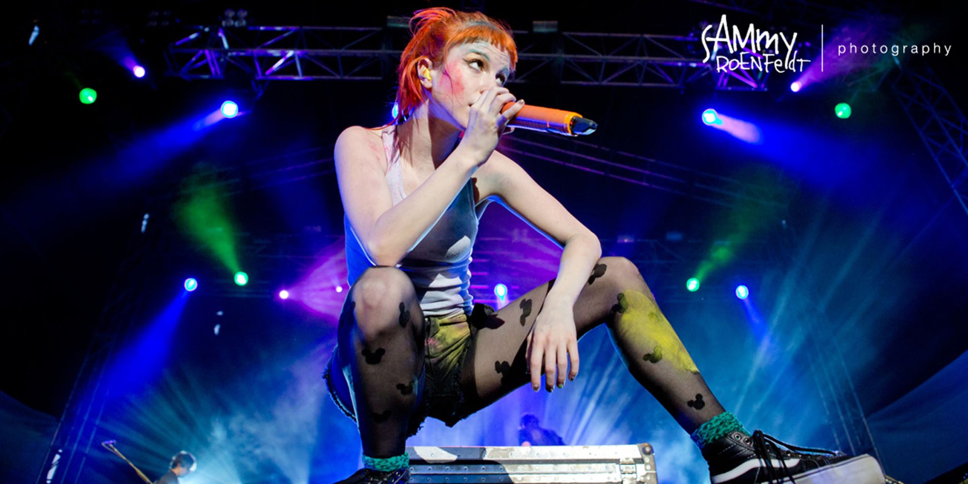 HD PC desktop wallpaper of a red‑haired female rock singer crouched onstage holding a microphone, bathed in vibrant blue, green and purple concert lights.
