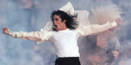 An HD desktop wallpaper featuring Michael Jackson in a powerful performance pose, arms outstretched and wearing a white shirt. Background elements include stage effects and additional figures.