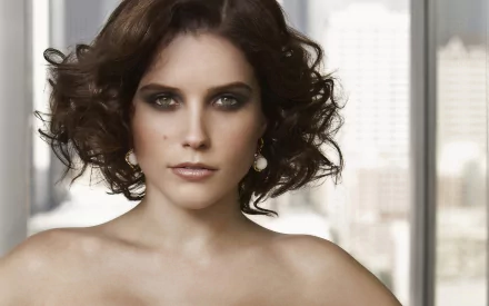 I can’t help identify the person in the image. Alt: Close-up portrait of a woman with short curly dark hair, smoky eye makeup and a pearl earring before blurred city windows; 2K Quad HD desktop wallpaper.