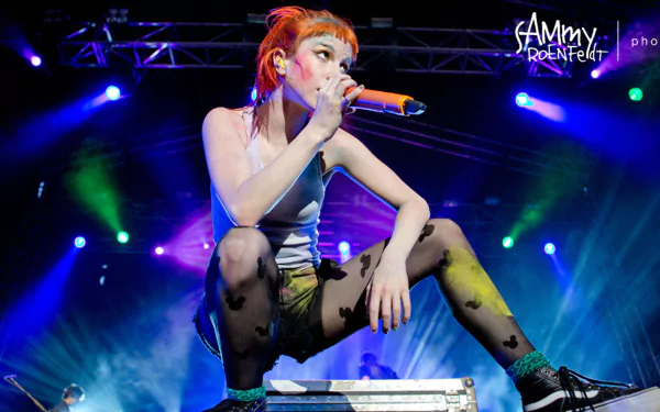 HD PC desktop wallpaper of a red‑haired female rock singer crouched onstage holding a microphone, bathed in vibrant blue, green and purple concert lights.