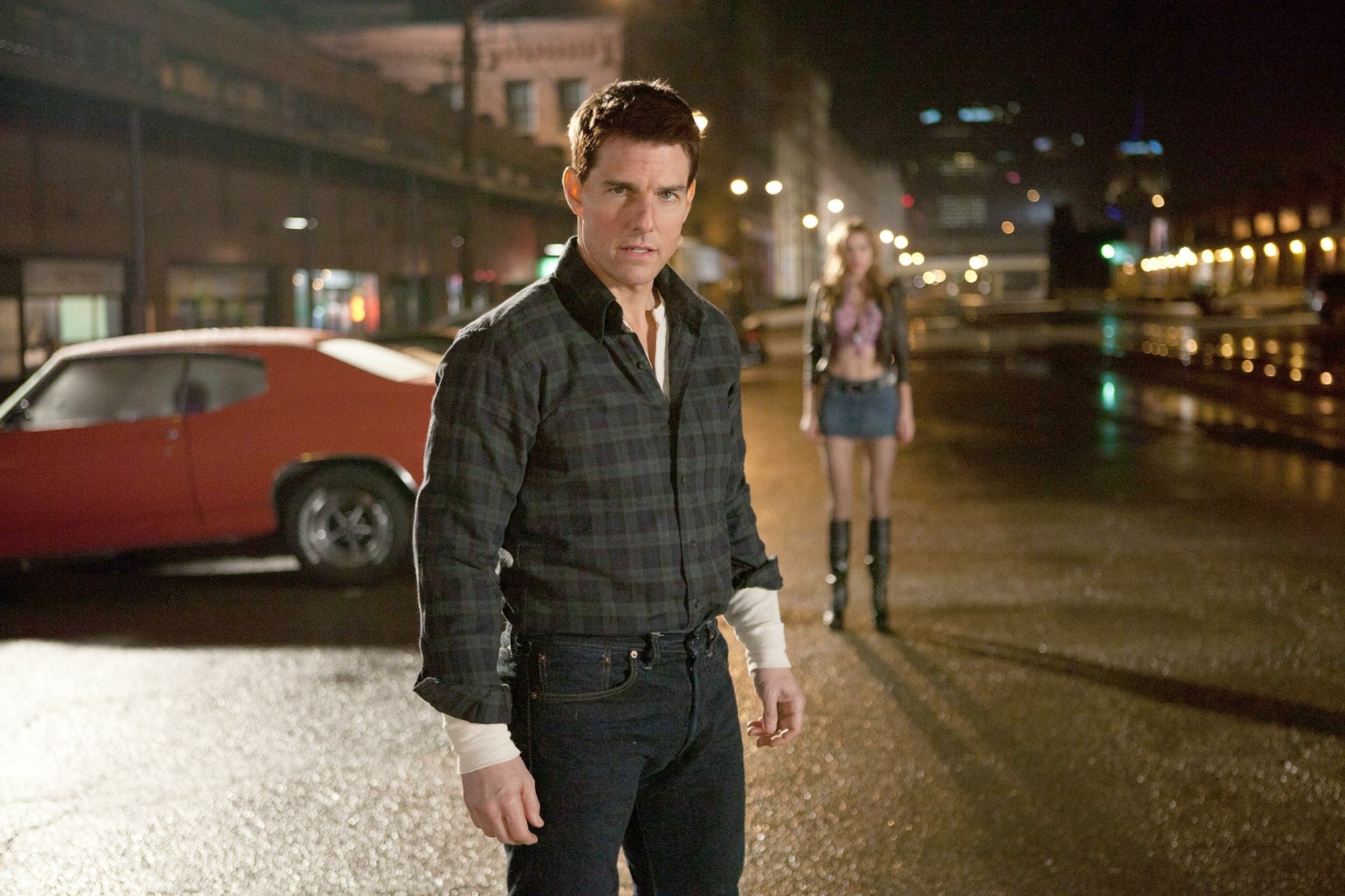 HD desktop wallpaper featuring Tom Cruise as Jack Reacher standing in a dimly lit street with a vintage car and a woman blurred in the background.