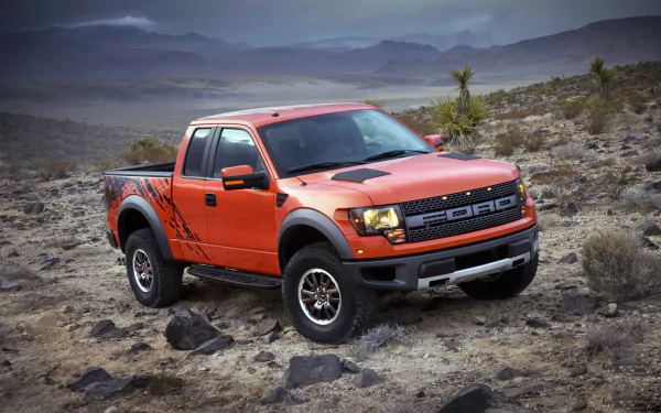 HD PC desktop wallpaper of an orange Ford F-150 Raptor pickup on rocky desert terrain, a rugged off-road vehicle set against distant mountains.