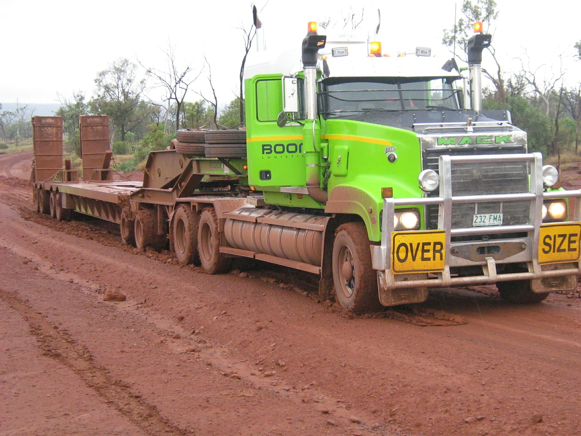 HD PC desktop wallpaper of a bright green Mack Trucks tractor towing a low-loader trailer along a muddy dirt road.