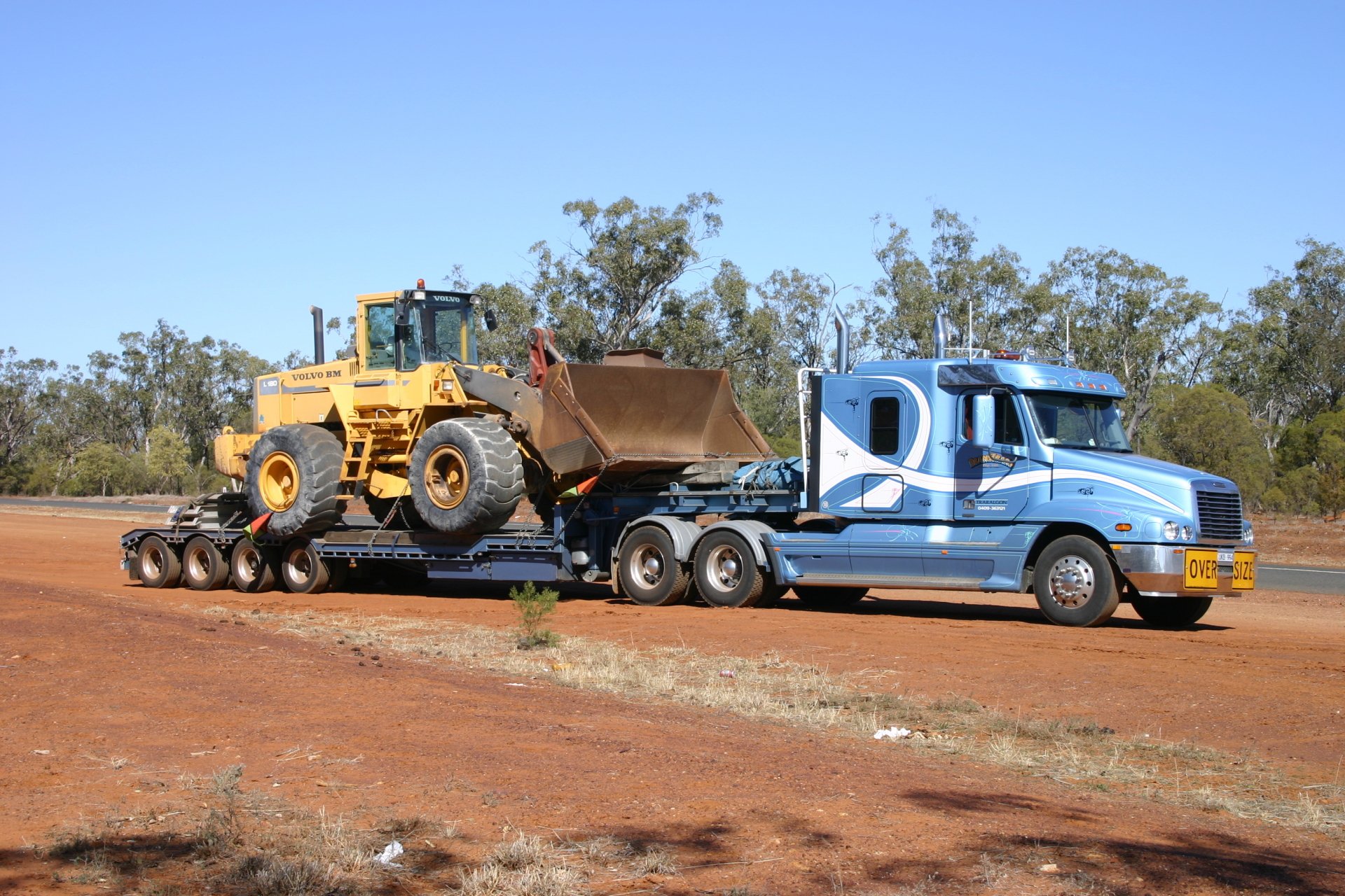 HD desktop wallpaper featuring a Freightliner truck transporting heavy construction equipment on a lowboy trailer against a clear sky and natural landscape backdrop.