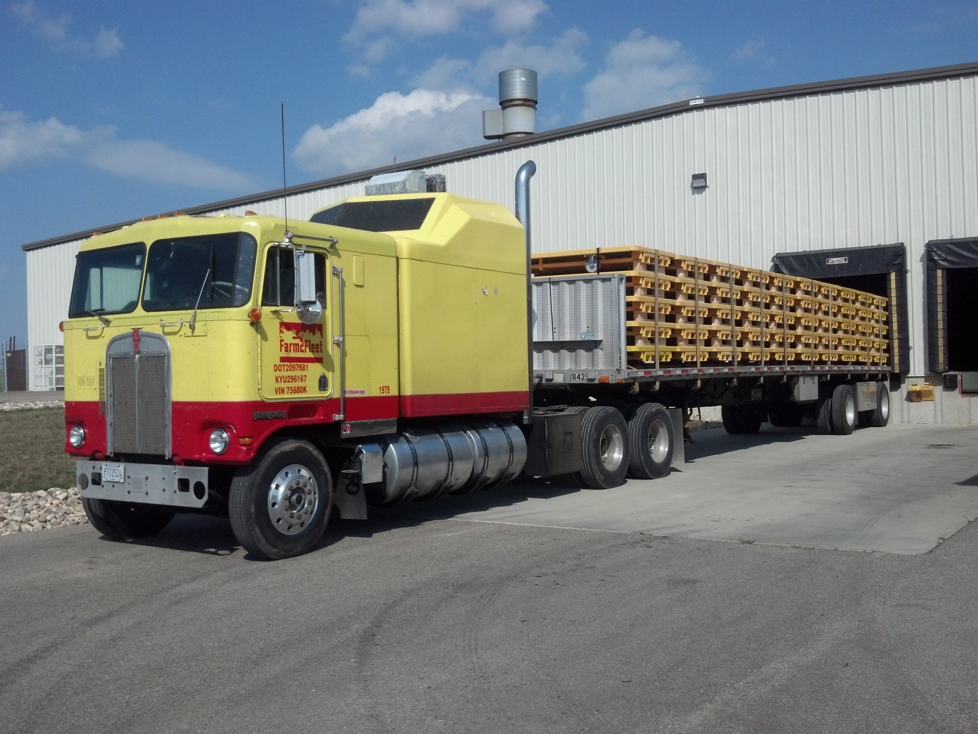 HD desktop wallpaper featuring a yellow and red Kenworth vehicle hauling a flatbed trailer loaded with lumber in front of a warehouse.