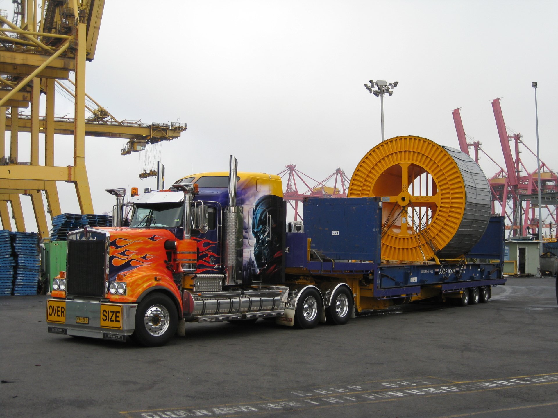 HD desktop wallpaper featuring a Kenworth vehicle transporting oversized industrial equipment at a busy port with cranes in the background.