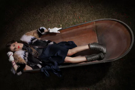 A stylish Alexandra Daddario reclines in a wooden wheelbarrow, wearing a chic outfit with fur accents and bold accessories, creating an eye-catching HD wallpaper.