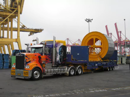 HD desktop wallpaper featuring a Kenworth vehicle transporting oversized industrial equipment at a busy port with cranes in the background.