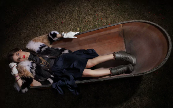 A stylish Alexandra Daddario reclines in a wooden wheelbarrow, wearing a chic outfit with fur accents and bold accessories, creating an eye-catching HD wallpaper.