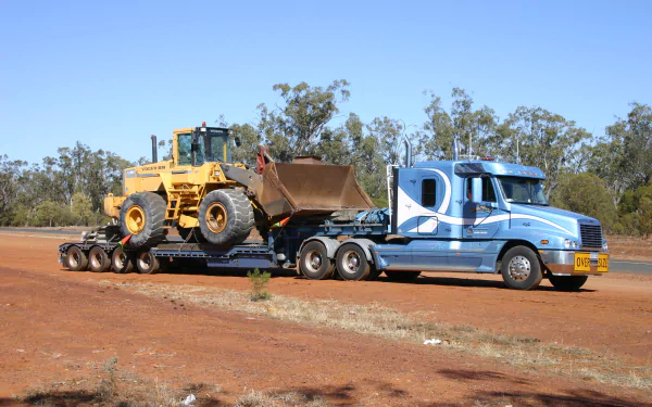HD desktop wallpaper featuring a Freightliner truck transporting heavy construction equipment on a lowboy trailer against a clear sky and natural landscape backdrop.
