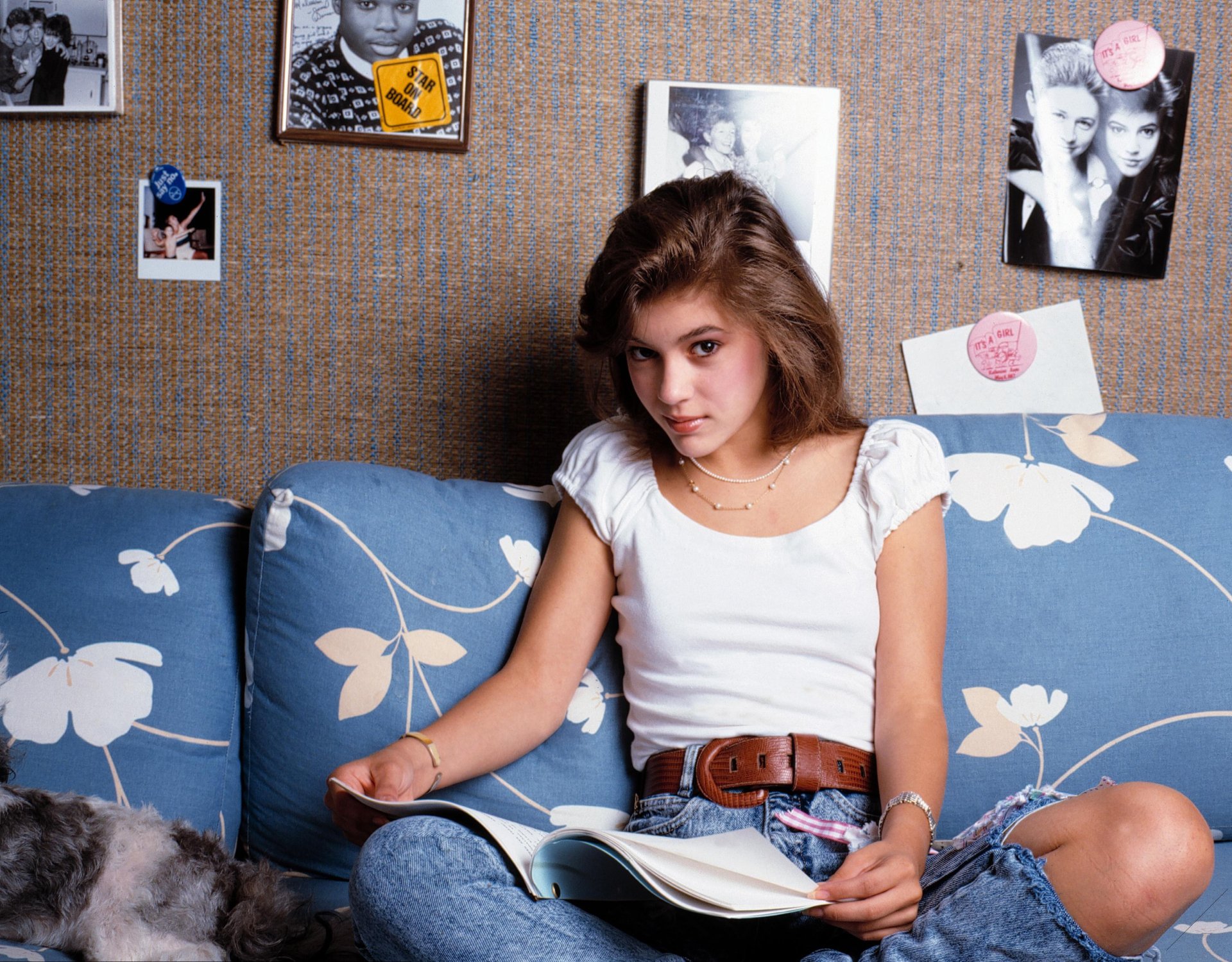 2K Quad HD PC wallpaper: young woman in a white top and jeans seated on a blue floral couch, reading a magazine against a poster-pinned wall, relaxed retro 1980s vibe.