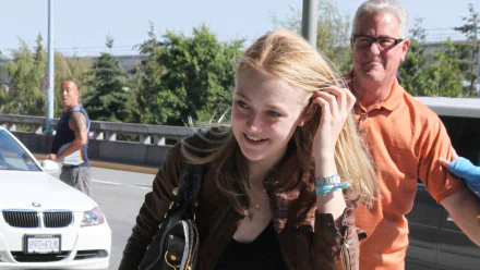 Blonde female celebrity smiling outdoors at an airport, walking past cars with a man in an orange shirt behind her — HD PC desktop wallpaper/background.