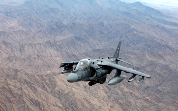 HD desktop wallpaper featuring a McDonnell Douglas AV-8B Harrier II military jet flying over a rugged desert mountain landscape.