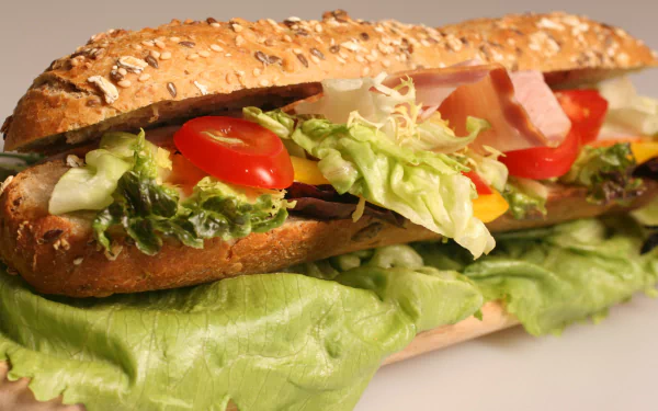 A close-up HD desktop wallpaper of a fresh sandwich with lettuce, tomato, and sliced deli meat inside a seeded bread roll, highlighting vibrant lunch ingredients.