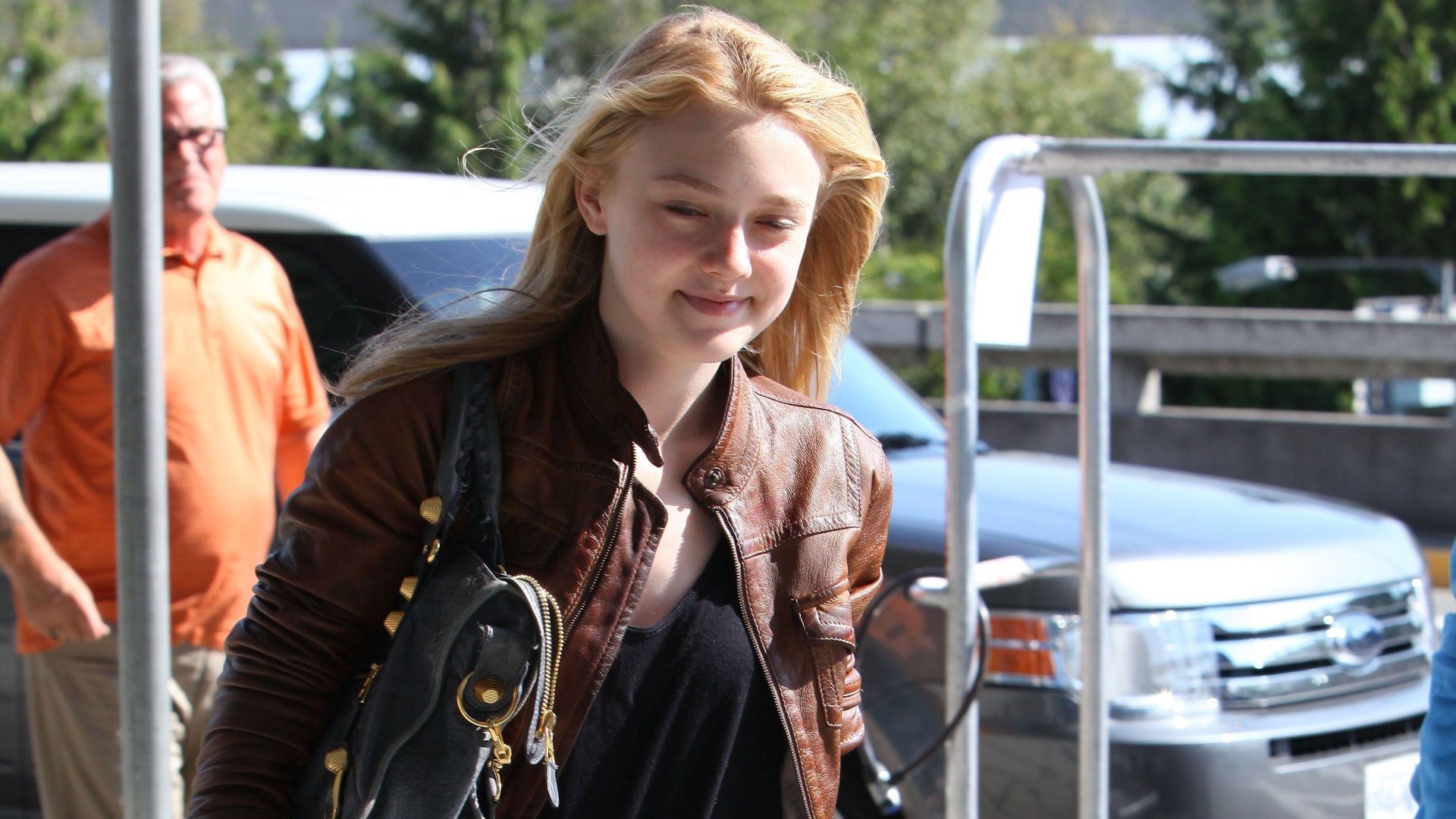 2K Quad HD PC desktop wallpaper of a young female celebrity smiling outdoors in a brown leather jacket, carrying a black handbag.