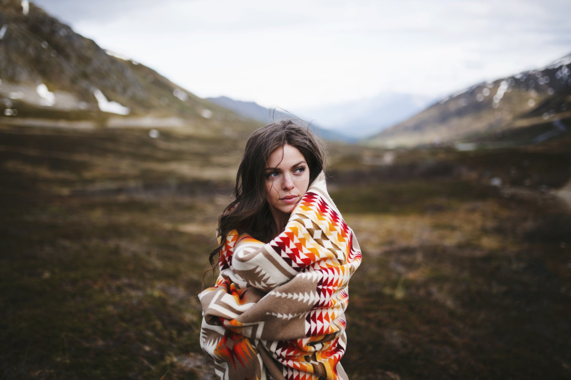 A brunette woman with blue eyes wrapped in a patterned blanket stands in a vast mountainous landscape, captured in crisp HD for a PC desktop wallpaper.