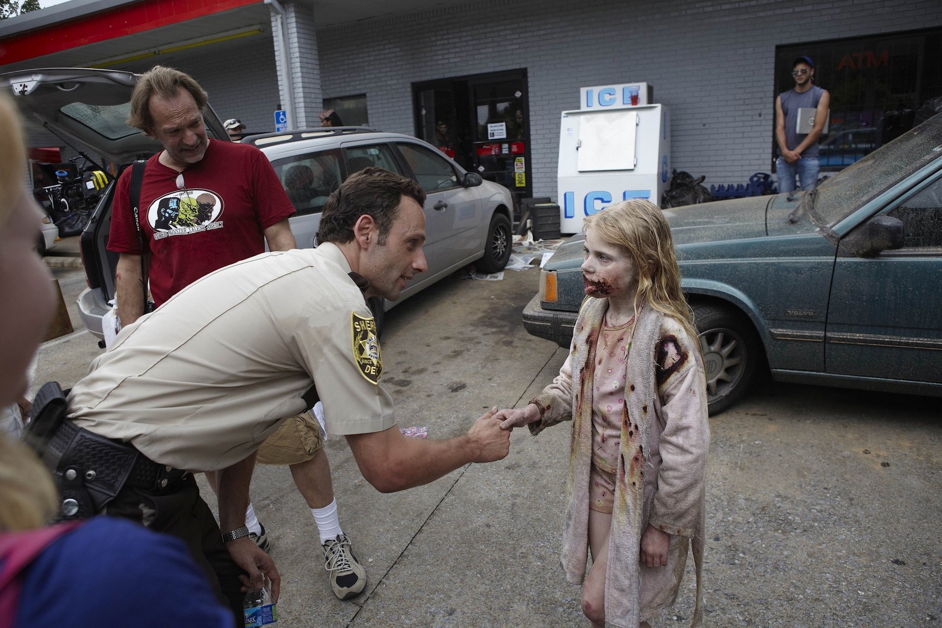 A scene from *The Walking Dead*, featuring Andrew Lincoln as a sheriff interacting with a young girl in a zombie costume, set against a backdrop of vehicles and a gas station.