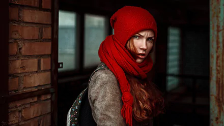 HD PC desktop wallpaper of a moody young woman in a red knit hat and scarf, intense gaze against a dim brick and windowed interior, warm autumnal tones.