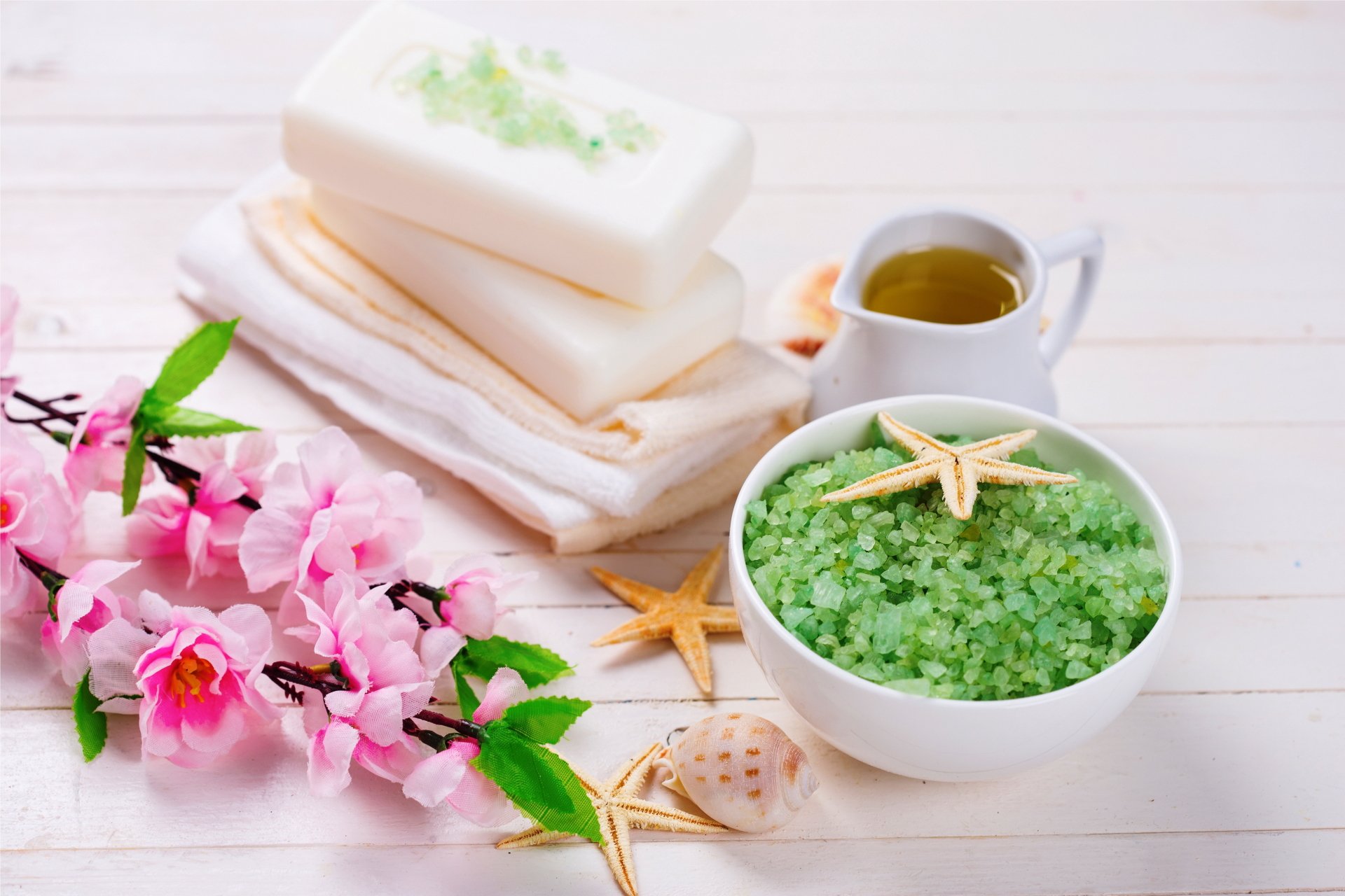 4K Ultra HD spa still life featuring green bath salt in a bowl with a starfish, pink flowers, white soap bars, a pitcher, and folded towels on a white wooden surface.