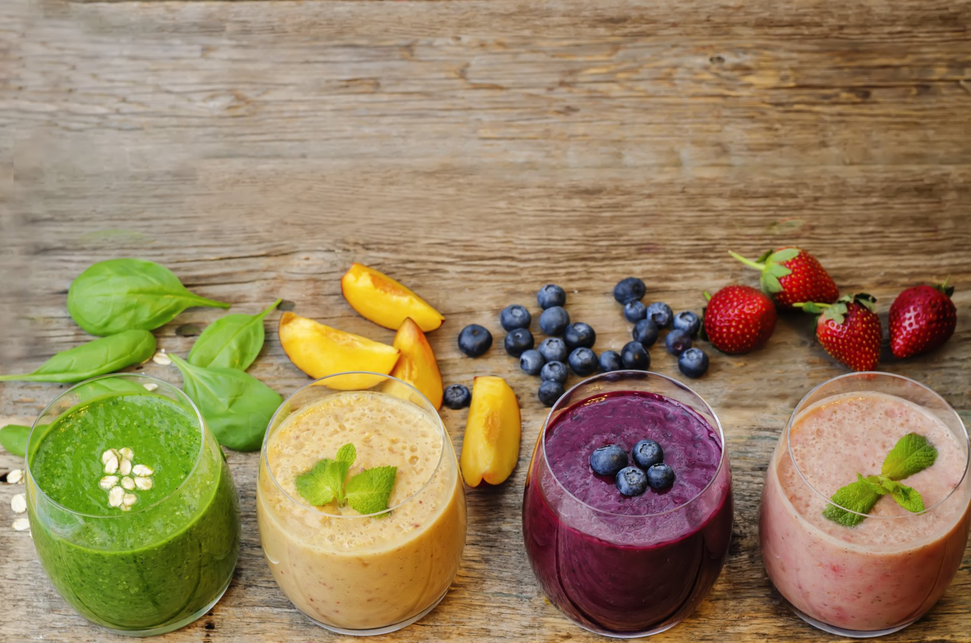 A vibrant 4K Ultra HD desktop wallpaper featuring four glasses of smoothies in green, peach, blueberry, and strawberry flavors, surrounded by fresh fruits and herbs on a wooden surface.