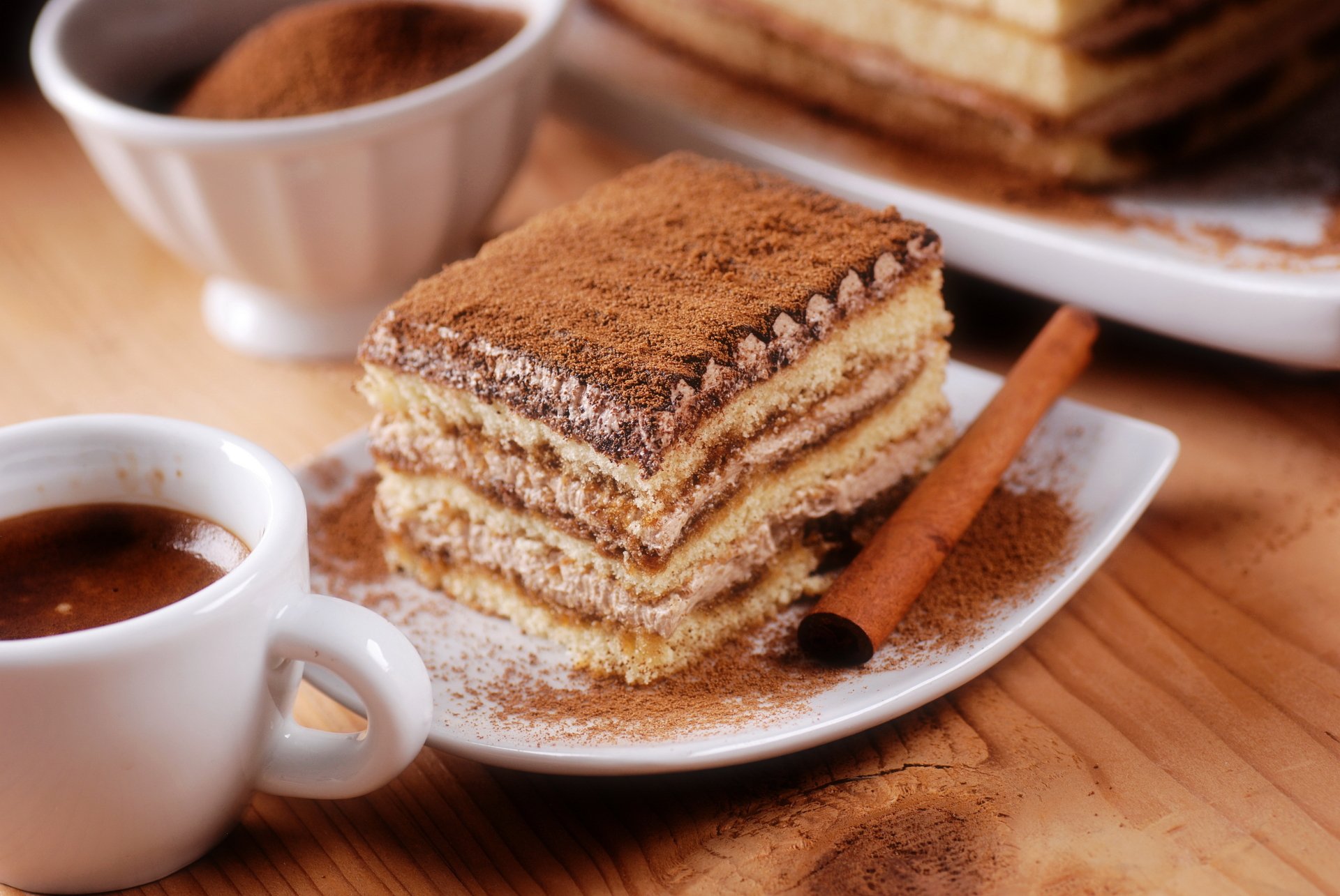 A close-up of a layered chocolate cake slice dusted with cinnamon, accompanied by a cinnamon stick, a cup of coffee, and a bowl of cinnamon powder, shown in 4K Ultra HD.