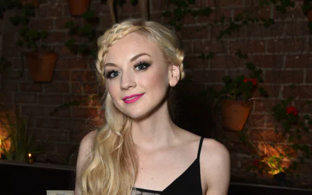 HD PC desktop wallpaper of American singer and actress Emily Kinney smiling against a dimly lit brick wall with potted plants in the background.