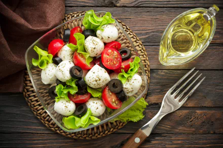 HD desktop wallpaper showing a fresh mozzarella, tomato, and olive salad with lettuce, served on a glass plate beside a bottle of olive oil and a fork on a wooden table.