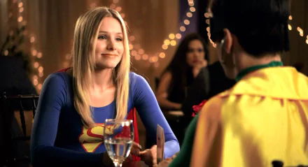 HD desktop wallpaper featuring Kristen Bell in a scene from the movie Movie 43, smiling and dressed in a blue Superman-themed outfit at a restaurant table.