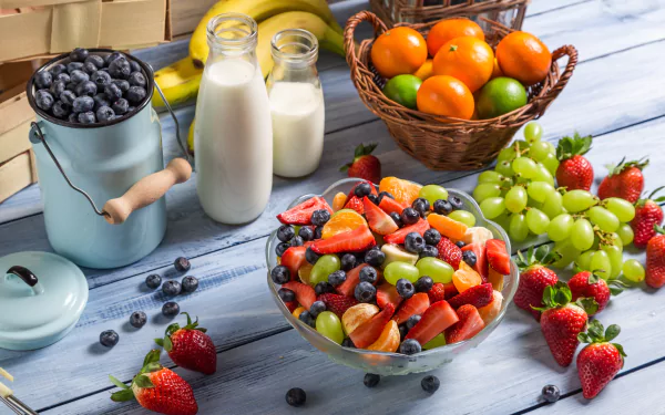 A vibrant arrangement of fresh fruit, including strawberries, blueberries, and grapes, alongside bottles of milk and a basket of oranges, set against a rustic wooden background.