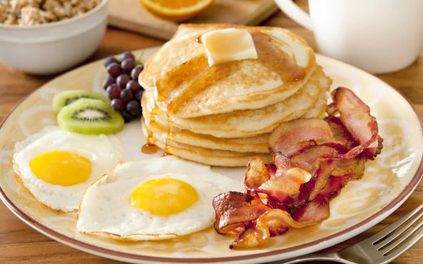 A delicious breakfast scene featuring pancakes topped with butter, crispy bacon, fried eggs, kiwi, and grapes, captured in vibrant 4K Ultra HD for a mouthwatering desktop wallpaper.