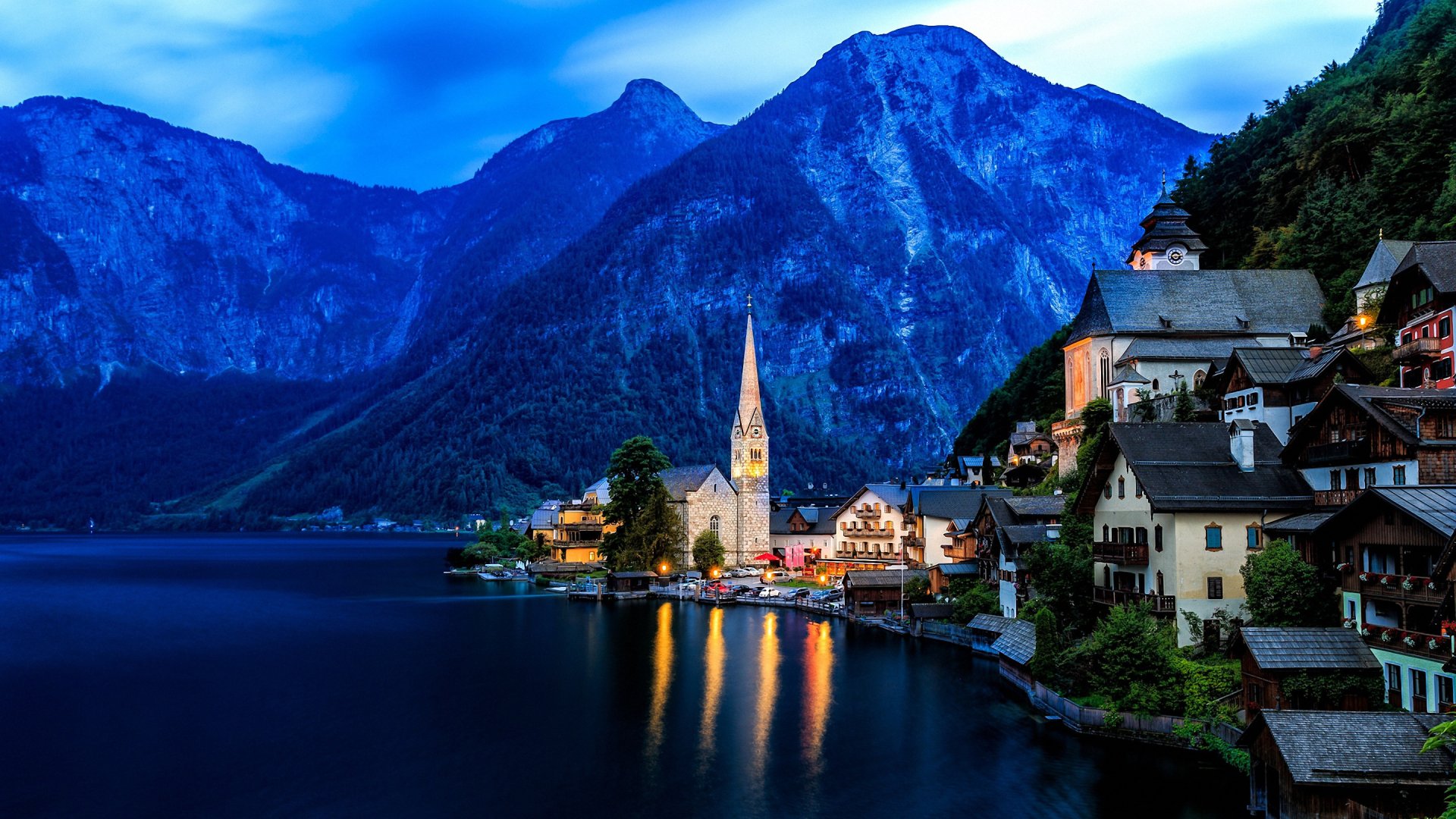 HD desktop wallpaper of Hallstatt village by a serene lake, surrounded by towering mountains and illuminated man-made lights reflecting on the water at dusk.