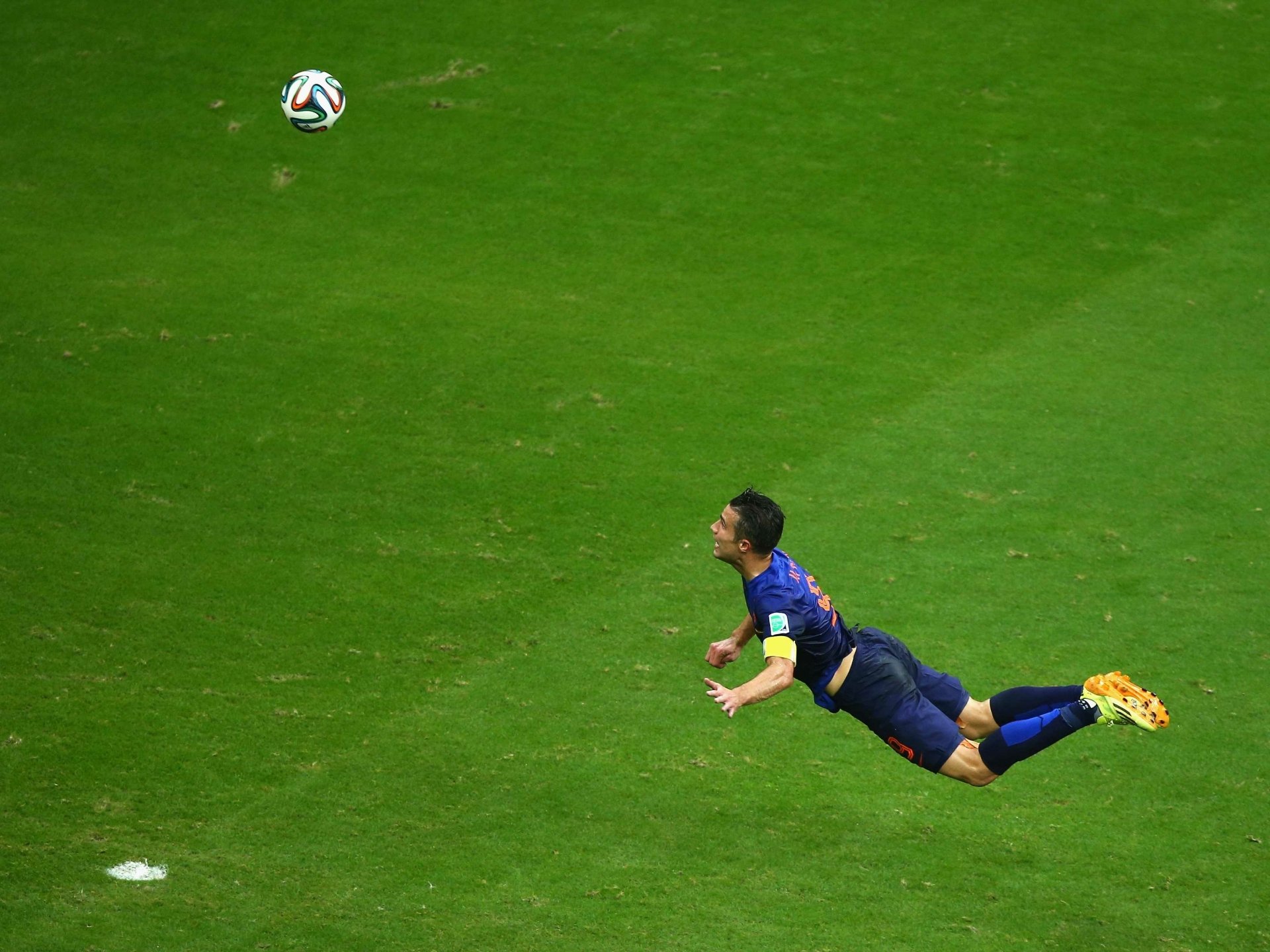 Diving soccer player midair heading a ball on a green pitch, sports action with captain's armband visible — 2K Quad HD PC desktop wallpaper and background.