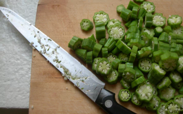 Close-up of sliced fresh okra with a chef's knife on a wooden cutting board, vibrant food image rendered as a 2K Quad HD PC desktop wallpaper and background.