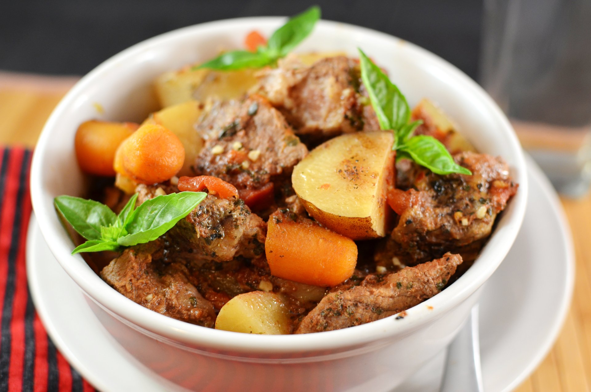 Close-up 4K Ultra HD image of a hearty stew with tender meat, carrots, potatoes, and fresh basil leaves in a white bowl on a wooden table.