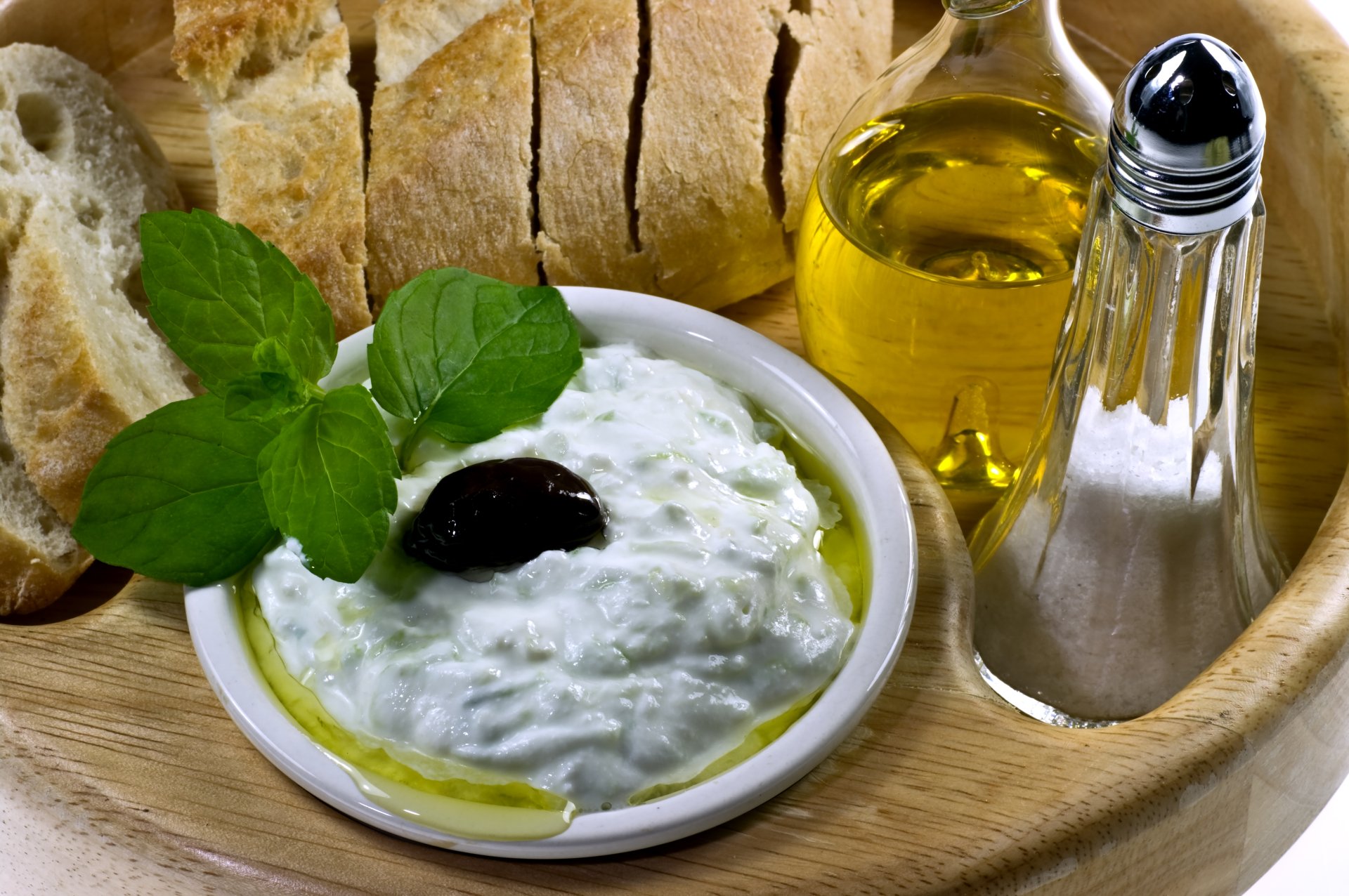 Close-up of a bowl of creamy tzatziki with olive oil, olive, fresh mint, sliced bread, and condiments, captured in 4K Ultra HD for a vivid PC desktop wallpaper.